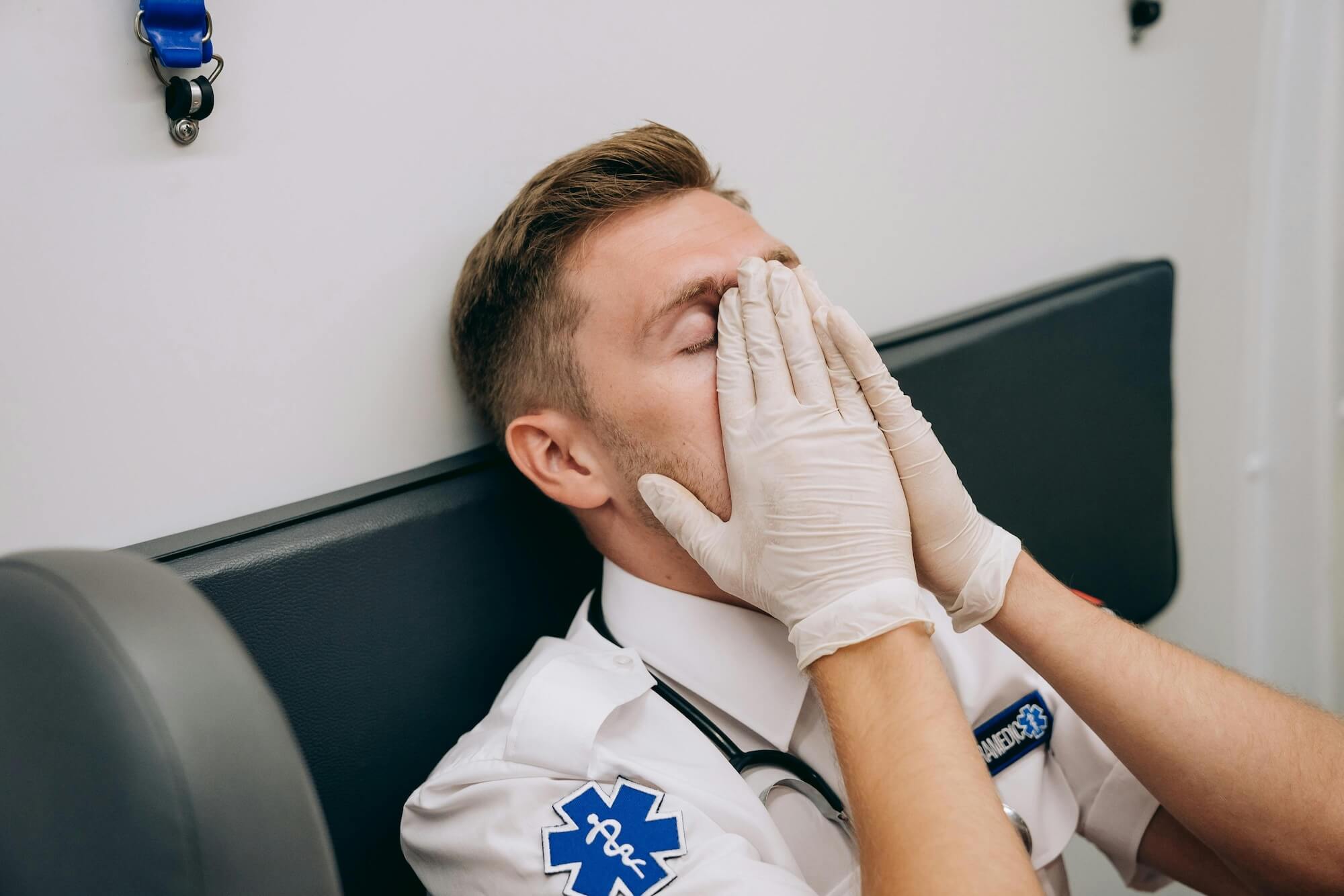 A paramedic wearing a uniform and medical gloves sits on a bench inside an ambulance, leaning forward with his hands covering his face, appearing tired or stressed.