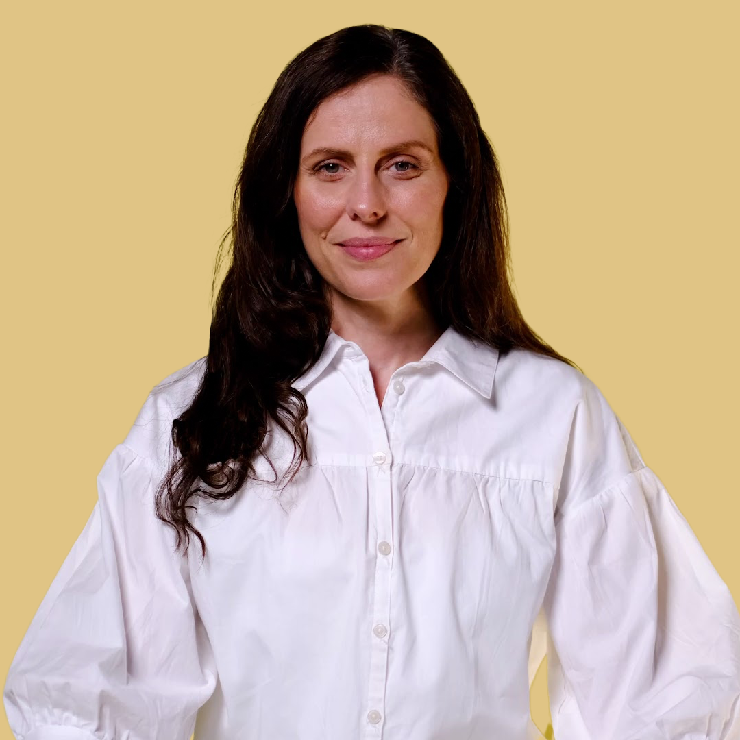 A woman with long dark hair wearing a white button-up shirt stands in front of a plain light tan background, looking at the camera with a slight smile.