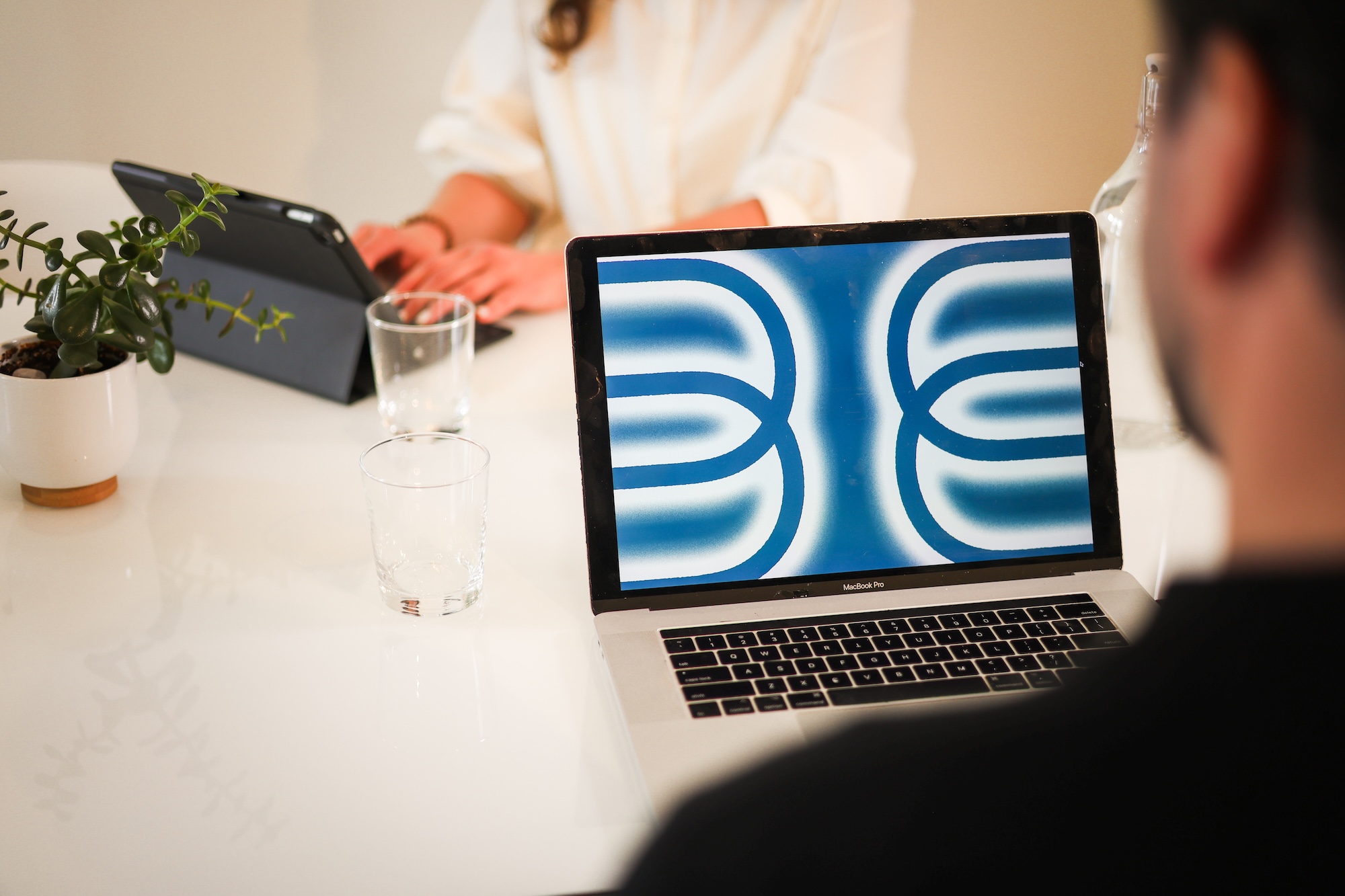 Two people sit at a table with laptops and glasses of water. The foreground laptop displays a blue and white abstract pattern. A small potted plant and a glass bottle are also on the white table.