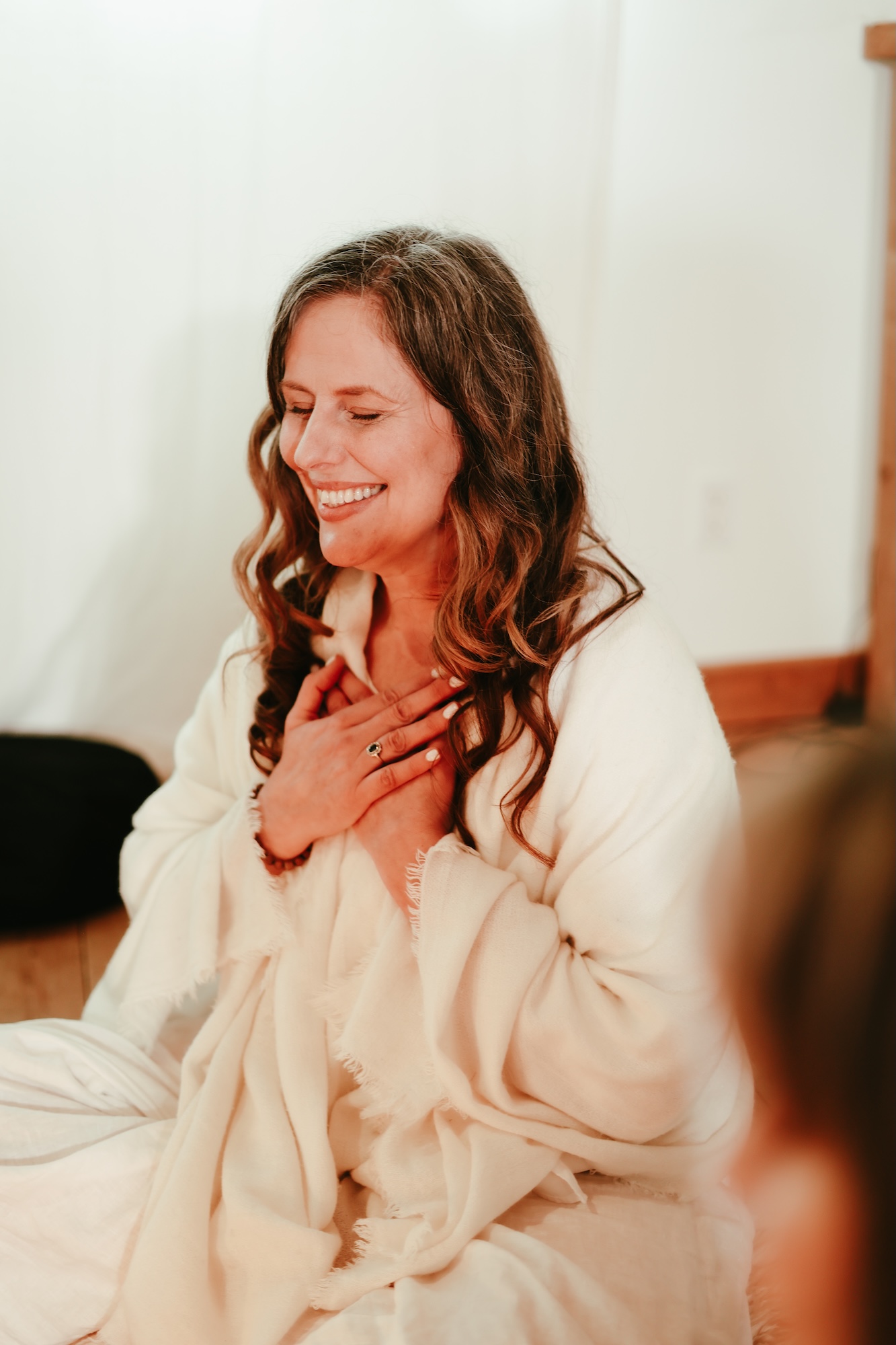 A woman with long brown hair sits indoors, smiling with her eyes closed and her hands placed on her chest. She is wearing a white shawl, and the background is softly lit.