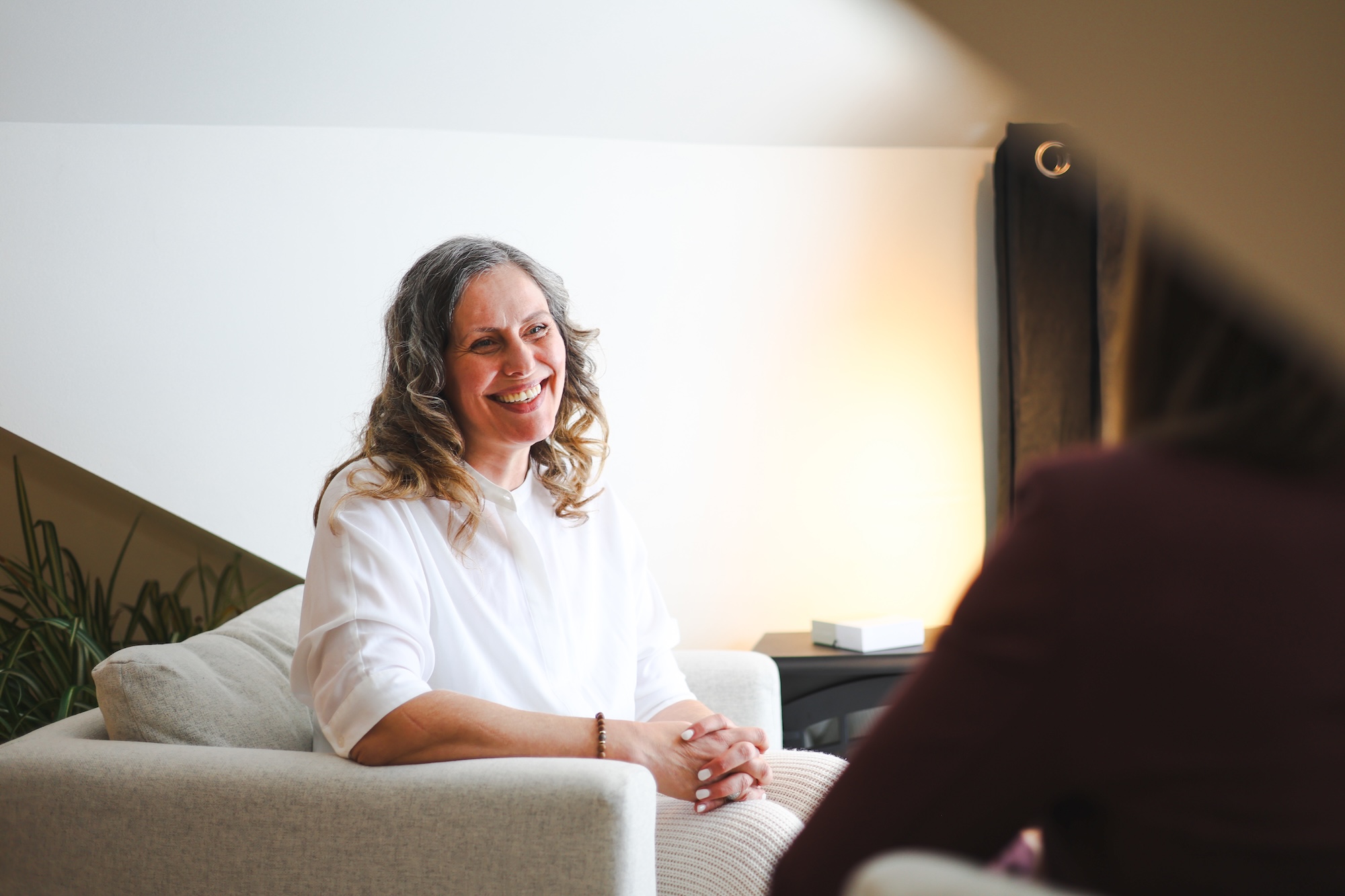 A woman with long, wavy hair and a white shirt sits on a light-colored armchair, smiling and talking to another person whose back is visible in the foreground. The room has soft lighting and light-colored walls.