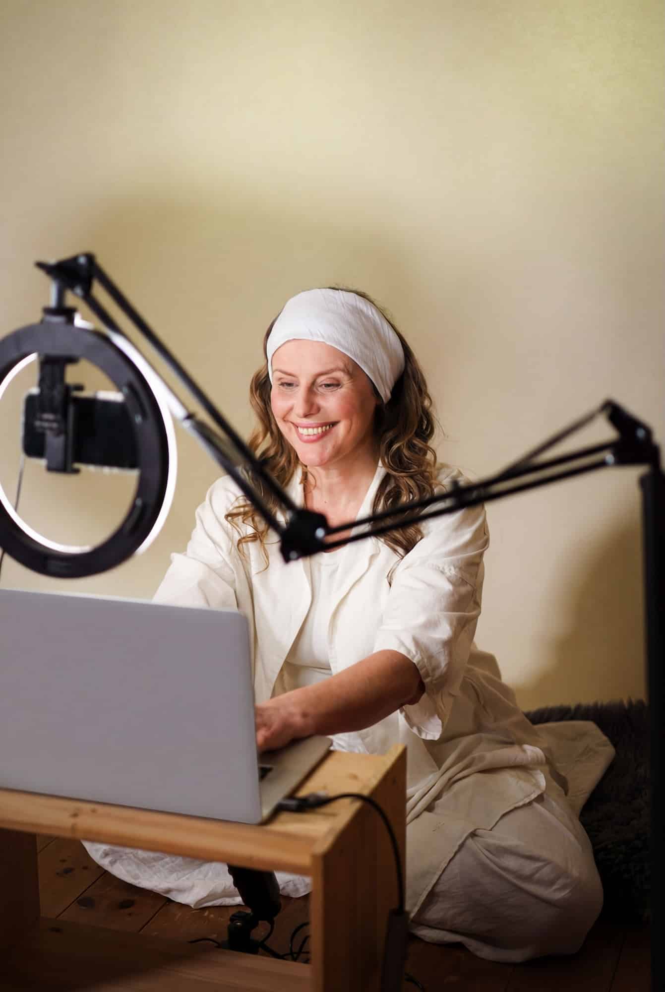 A woman wearing a white outfit and headband sits on the floor, smiling at a laptop. She is surrounded by a ring light and a microphone, suggesting she is recording or live streaming content.