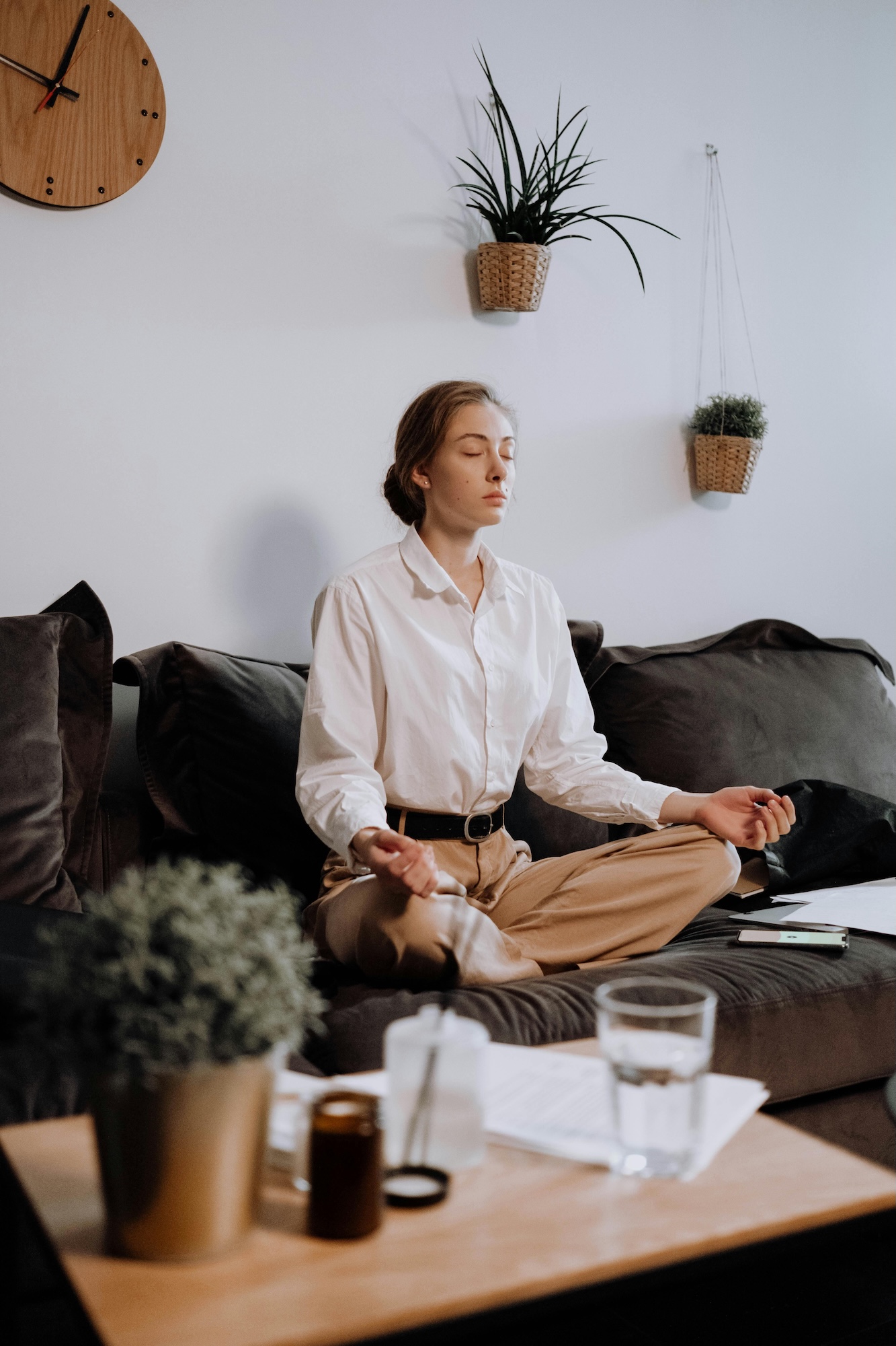 A woman in business attire sits cross-legged on a couch with her eyes closed, meditating. A table with a glass of water and potted plant is in front of her. Wall-mounted planters and a wooden clock are in the background.