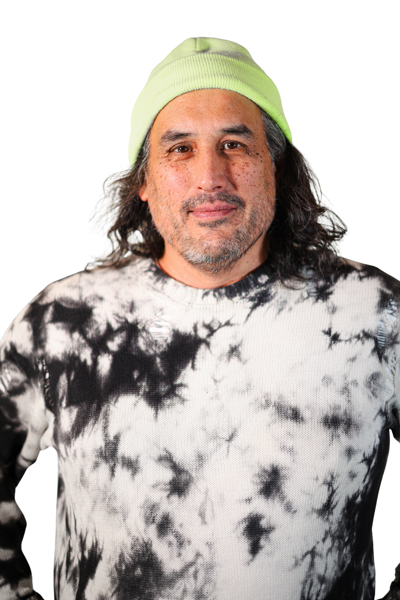 A person with long dark hair and a beard, wearing a light green beanie and a black and white tie-dye sweater, stands against a plain white background.
