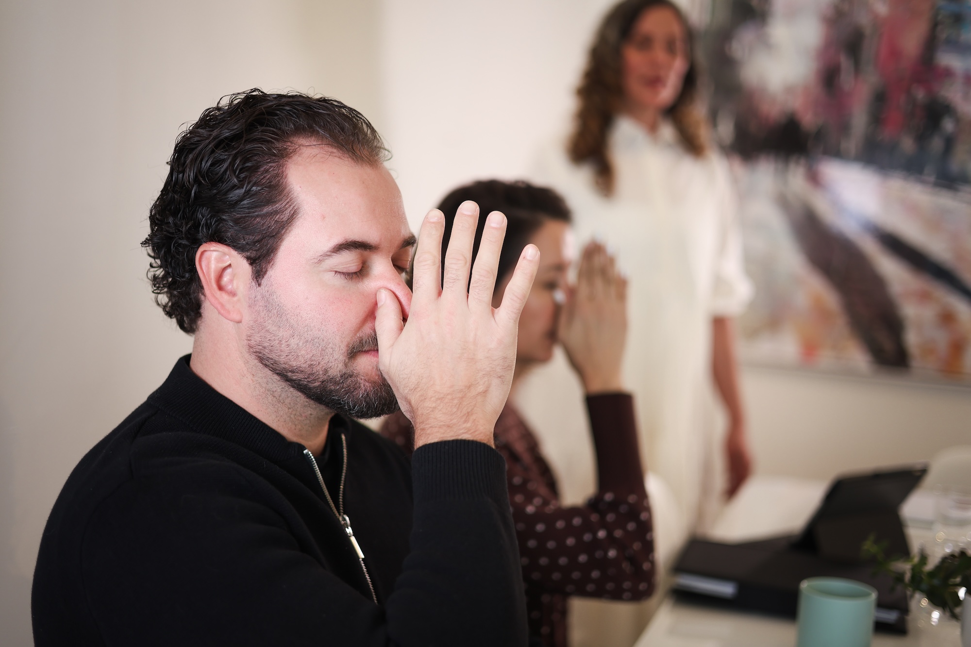 Three people sit at a table; two in the foreground are practicing a breathing exercise with fingers on their noses, while a woman in the background stands and observes.