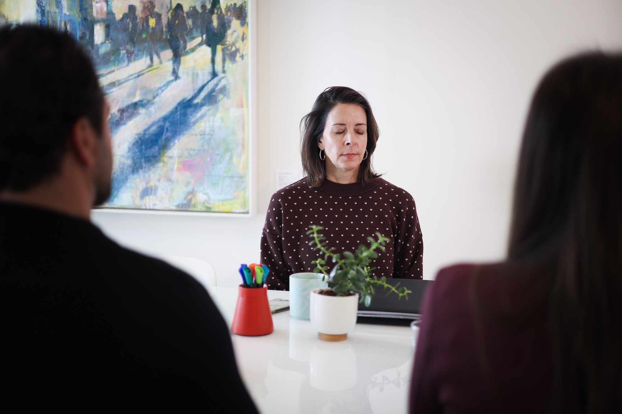 Three people sit at a table; two in the foreground are practicing a breathing exercise with fingers on their noses, while a woman in the background stands and observes.