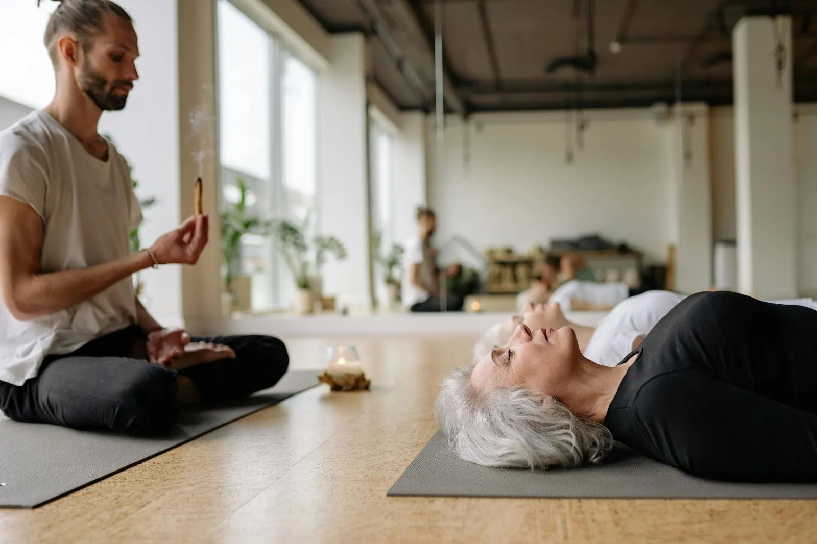 A person sits cross-legged holding incense while another person lies on a yoga mat with eyes closed in a bright, spacious room with large windows and indoor plants.